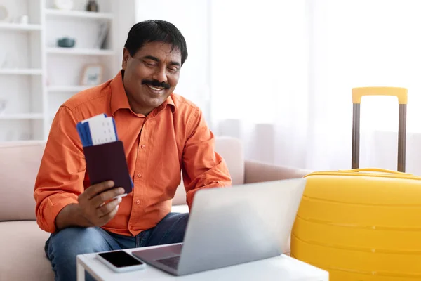 Online tickets booking. Excited mature indian man using laptop and buying tour on internet travel agency, posing near suitcase at modern home interior, holding passport. Vacation plans concept