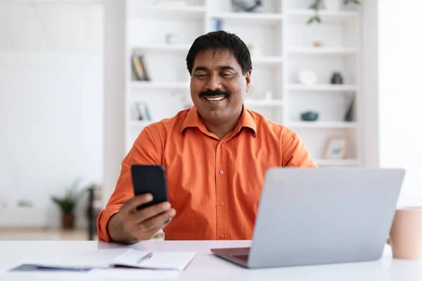 Multitasking at work concept. Positive middle aged hindu businessman using smartphone and laptop computer at home office, replying emails, websurfing. Digital world and business concept