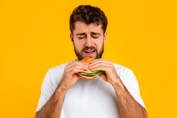 Unhappy Bearded Young Man Eats Burger Portraying Emotional Struggle Balancing — Stock Photo, Image