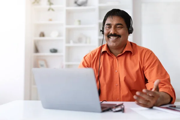 Remote work, freelance. Cheerful middle aged eastern man with moustache sitting at desk, using laptop computer and headset, have business meeting, working from home office, copy space