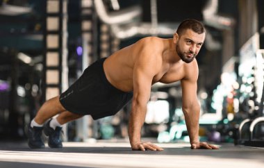 Spor salonunda antrenman yaparken odaklanmış, kaslı erkek sporcu yoğunluğunu ve gücünü ifade ediyor, modern spor kulübünün iç kısımlarında spor yapan, kalas üzerinde duran üstsüz adam.