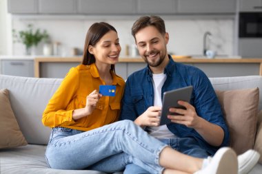 Neşeli genç çift tabletle takılıyor, kredi kartı tutan kadın online alışveriş veya rezervasyon yaptıklarını öne sürüyor, kanepede oturuyorlar.