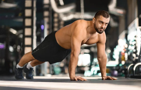 Focused man doing mid-push-up exercise while training at gym, muscular male athlete expressing intensity and strength, shirtless guy exercising in modern sport club interior, standing in plank
