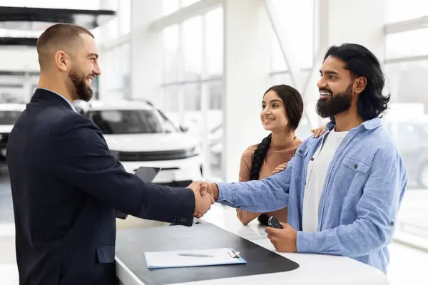 Side view of auto salesman wearing suit shaking happy indian guy customer hand, manager and spouses standing at counter. Cheerful young indian couple buying new car at dealership salon