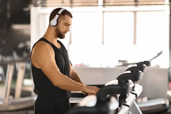 Jogging Workout. Portrait Of Young Muscular Man Wearing Wireless Headphones Using Treadmill At Gym, Motivated Male Athlete Exercising In Modern Fitness Club, Enjoying Active Lifestyle, Copy Space