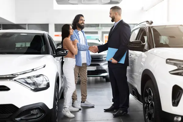 With warm smile and clipboard in hand, a professional car advisor stands discussing financing options with an interested indian couple at a dealership, shaking man client hand, full length