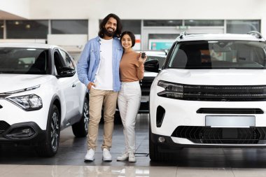Gülümseyen milenyum Hintli çift yeni beyaz arabanın yanında duruyor, yepyeni lüks otomobilin anahtarlarını kucaklıyor ve gösteriyorlar, oto galerisinde alışverişi kutluyorlar, tam boy