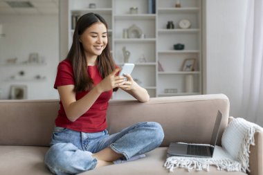 Evde akıllı telefon ve dizüstü bilgisayar kullanan gülümseyen Asyalı genç bir kadın bilgisayarla uzaktan çalışmaktan, oturma odasında kanepede oturmaktan, uzay kopyalamaktan mutlu Koreli bir kadın.