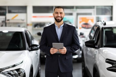 Gülümseyen, takım elbiseli yakışıklı adam araba galerisinde duruyor ve dijital tablet kullanarak yeni mesajları kontrol ediyor müşteriler internete yüklüyor, kameraya gülümsüyor.