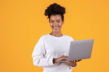 Dizüstü bilgisayarı tutan ve kameraya gülümseyen kendinden emin genç siyahi kadın. Elinde bilgisayarla, parlak sarı arka planda izole bir şekilde poz veren, mutlu, Afro-Amerikan bayan.