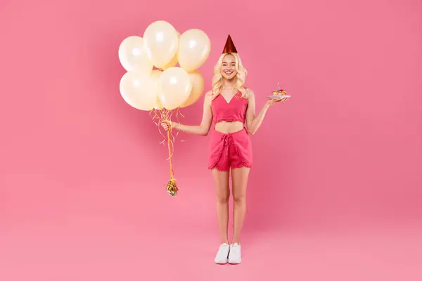 お祝いの帽子で陽気な若いブロンドの女性 クリーム風船と誕生日ケーキを保持 活気に満ちたピンクの背景に対して遊び心のあるポーズと立って — ストック写真
