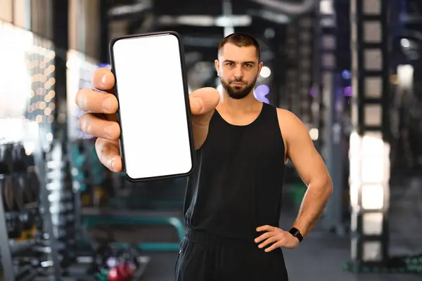 Fitness App. Sporty Young Man Showing Smartphone With Blank White Screen At Camera, Handsome Muscular Male Athlete Recommending New Mobile Application For Workout Trainings, Posing At Gym, Mockup
