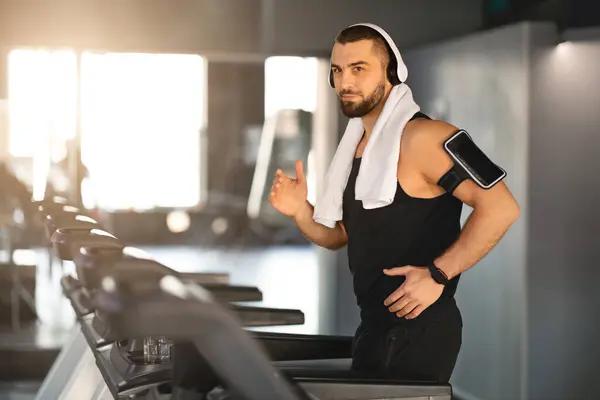 Muscular Male Athlete Wearing Wireless Headphones Running On Treadmill At Gym, Motivated Millennial Man Listening Music During Workout, Sporty Guy Enjoying Cardio Training, Copy Space