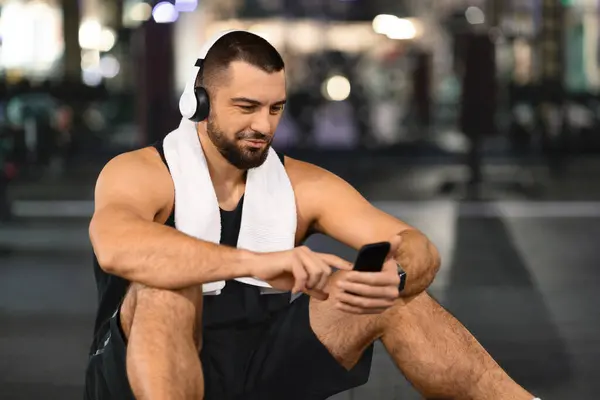 Handsome Young Athlete Man Relaxing With Smartphone After Training In Gym, Smiling Muscular Guy Wearing Wireless Headphones Sitting On Floor And Listening Favorite Music On Mobile Phone, Copy Space