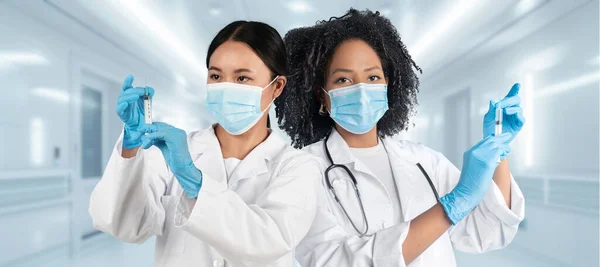 Two diverse european, african american women healthcare workers in white coats and blue gloves hold syringes while wearing surgical masks in a clinical hospital setting, panorama
