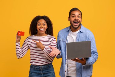 Kolay online bankacılık konsepti. Mutlu siyah çift dizüstü bilgisayar kullanıyor ve kredi kartını işaret ediyor. Heyecanlı Afrikalı Amerikalı eşler sarı stüdyo arka planında poz veriyorlar. Bilgisayarla online alışveriş yapıyorlar.