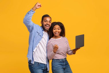 Hevesli genç siyah çift ellerinde dizüstü bilgisayarla başarıyı kutluyorlar, neşeli Afro-Amerikan erkek ve kadın, parlak sarı arka planda duruyorlar, başarı ve neşeyi somutlaştırıyorlar.