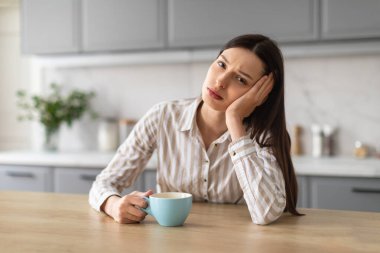 Endişeli, çizgili gömlekli genç bir kadın mutfak masasında oturuyor, elinde mavi bardakla sabahları kahve içiyor, üzgün bir ifadeyle kameraya bakıyor.