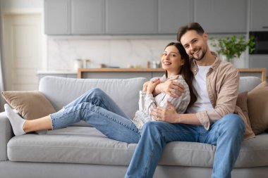 Neşeli genç çift rahat anın tadını çıkarıyor, erkek kadını arkadan kucaklıyor rahat kanepede rahatlarken, mutluluk ve neşe saçıyor.