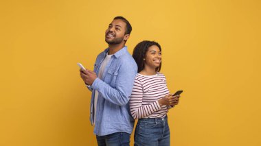 Hayalperest siyah çift sırt sırta durup cep telefonlarını tutuyorlar. Afro-Amerikan erkek ve kadın akıllı telefon kullanıyor ve sarı stüdyo arka planında fotokopi odasına bakıyorlar.
