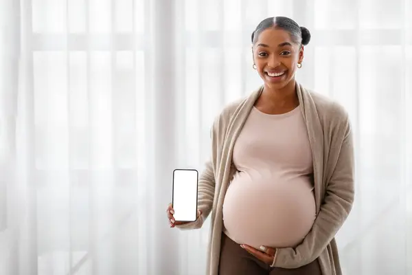 快乐的年轻怀孕黑人妇女展示手机 白色的空白屏幕模仿复制空间 站在窗边 抱着肚子 申请未来的妈妈 — 图库照片