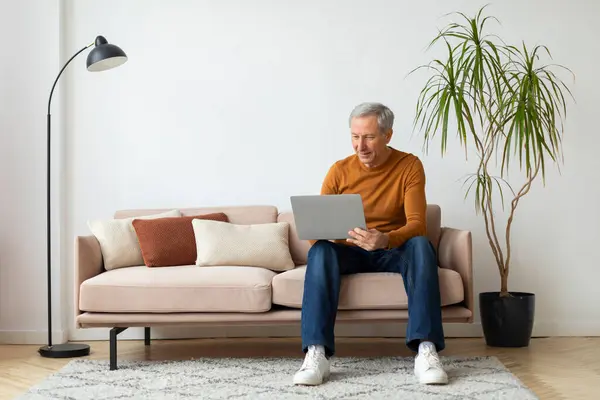 Handsome european old man using computer laptop at home, sitting on couch in cozy living room interior, looking for nice online course for seniors, searching good movie, copy space.