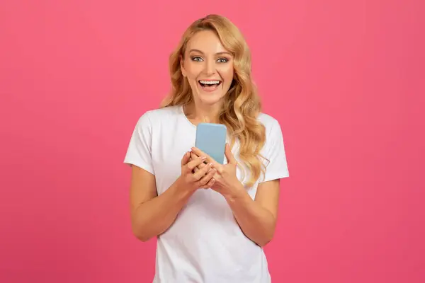 Great mobile offer. Joyful blonde millennial lady scrolling through smartphone, smiling to camera, texting in online chat on pink studio background. Social media apps, modern communication