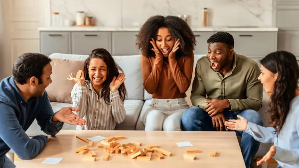 Diverse group of friends playing board game in living room, reacting emotionally as tower of blocks falling, creating moments of excitement and shared joy during indoor students gathering