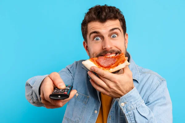 Emotional young European man enjoys pizza slice pointing remote controller at camera, watching favorite TV show and binge eating junk food, over blue studio background, closeup portrait