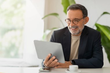 Gülümseyen Avrupalı yaşlı beyefendi ışık dolu bir ortamda tabletle etkileşime giriyor, ofiste çalışıyor, sohbet ediyor ve haber okuyor. Toplantı ve iletişim, iş görüşmesi, video görüşmesi.