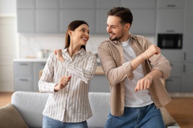 Genç eşler gündelik kıyafetleriyle dans ediyor ve rahat bir evde birlikte vakit geçiriyorlar. Birbirlerine bakıp gülümsüyorlar.