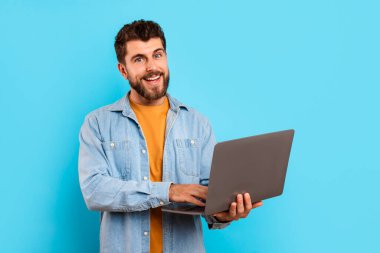 Neşeli bir milenyum adamı dizüstü bilgisayarda internette geziniyor, mavi arka planda duruyor, kameraya gülümsüyor. Modern profesyonel çevrimiçi çalışıyor. Teknoloji ve yazılım, internet işi