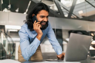 Ofiste çalışan neşeli gülümseyen genç Hintli bilişim müdürü, resmi olarak masa başında duran, dizüstü bilgisayarı kullanan ve iş ortağıyla telefon görüşmesi yapan,