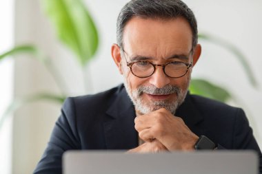 Bir dizüstü bilgisayarda çalışan, ofiste çalışan, ofiste haber okuyan ve video izleyen gülümseyen Avrupalı kıdemli bir adam. Toplantı ve iletişim, iş görüşmesi, video görüşmesi.