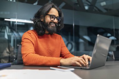 Başarılı sakallı Hintli adam proje yöneticisi gözlük takıyor ofiste çalışma masasında oturuyor, dizüstü bilgisayarda daktilo yazıyor ve gülümsüyor, müşterilerle sohbet ediyor, işinden zevk alıyor, boşluğu kopyalıyor.