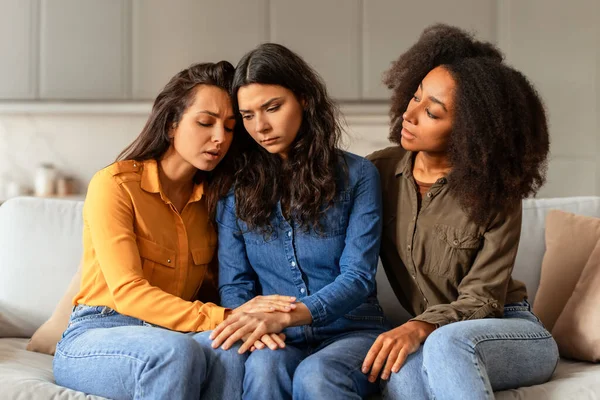 Compassionate diverse girlfriends sitting together providing support to unhappy woman friend going through life challenges and issues, sitting on couch in modern living room at home
