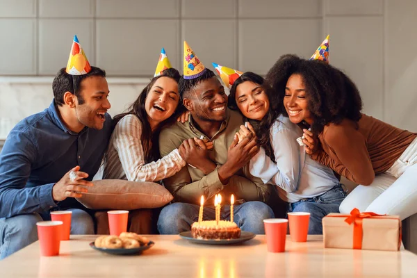 Diverse friends embracing birthday guy sitting with party hats, sharing joy and laughter in heartwarming celebration indoors. Group of multiracial students enjoying bday gathering together