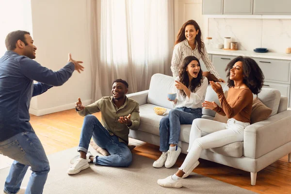 International group of millennials students gathering in living room for fun filled game of charades. Diverse young men and women laughing and engaging in entertaining activity during home party