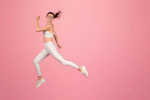 Energetic european young woman in motion, joyfully running in mid-air with a white sports bra and leggings against a pink backdrop, portraying vitality and an active lifestyle