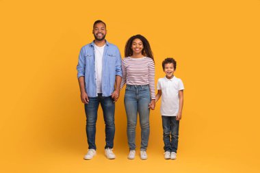 Gülümseyen siyahi ailenin küçük oğluyla el ele tutuşurken canlı sarı arka plan karşısında mutlu, Afro-Amerikan ebeveynler ve erkek çocuk stüdyoda poz verirken çekilmiş portresi.