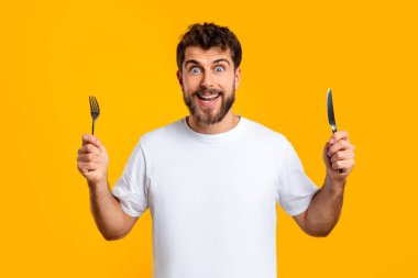 Açlık aşermeleri. Aç milenyum adamı elinde çatal bıçak tutuyor, lezzetli yemek beklentisi içinde, sarı stüdyo arkaplanının üzerinde günlük beyaz tişörtle fotoğraf makinesine gülümsüyor.