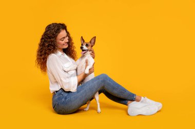 Kıvırcık saçlı genç kadın Jack Russell Terrier 'ını nazikçe tutuyor. İkisi de neşeli sarı arka plana karşı poz veriyor.