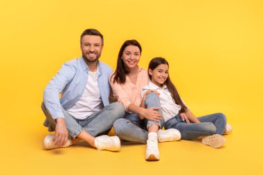 Sevgi dolu aile, sarı arka plan üzerinde yakın poz verir. Stüdyo çekimi. Mutlu anne, baba ve kız birlikte oturuyor ve gülümsüyor, mutluluğu ve aile bağını ifade ediyor.