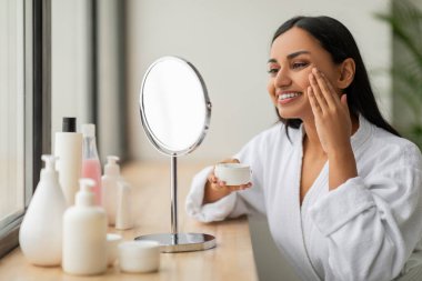 Cilt bakımı rutini. Beyaz bornozlu mutlu ve çekici bir Hintli kadın evde yüz kremi kullanıyor. Güzel bir bayan en yeni organik güzellik ürününü test ediyor. Makyaj masasında oturuyor.