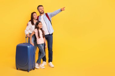 Üç kişilik bir aile, bavulları tatile hazır. Stüdyonun sarı arka planına karşı seyahat teklifleriniz için mutlulukla uzayı gösteriyorlar. Mutlu baba, anne ve kız bavulları fotokopi alanı gösteriyor.