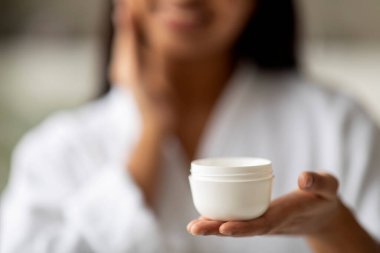 Yüz kremi ile el, cilt şımartma konsepti. Beyaz bornoz giyen, elinde nemlendirici tutan, duştan sonra yaşlanma önleyici krem kullanan doğulu bir kadın.