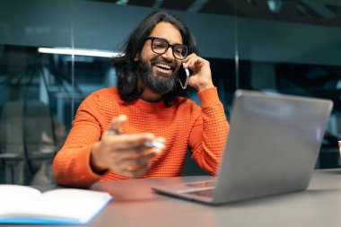 Turuncu kazaklı ve gözlüklü Hintli pozitif sakallı adam modern ofiste çalışıyor, masada oturuyor, dizüstü bilgisayar ekranına bakıyor ve telefonla konuşuyor.