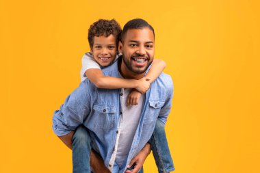 Mutlu siyah baba ve oğul, sarı stüdyo geçmişine karşı yürekten kucaklaşırlar, neşeli genç baba, neşeli ergen erkek çocuğunu sırtında taşır, her ikisi de neşeyle gülümserler, kopyalama alanı.
