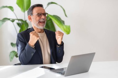 Gözlüklü kıdemli Kafkas işadamı dizüstü bilgisayarın önündeki yumruklarıyla başarıyı kutluyor, yemyeşil bitkilerle dolu modern bir ofiste zaferin tadını çıkarıyor.