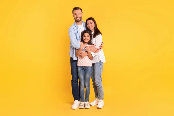 欢愉的欧洲家庭在随意拥抱的同时带着微笑看着相机 在黄色的工作室背景上摆出姿势 分享着爱的瞬间 全长的快乐父母和女儿 复制空间 — 图库照片